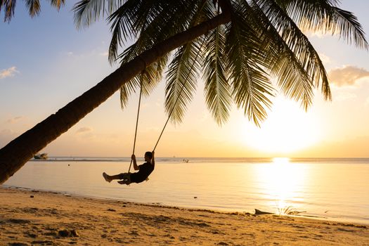 The guy enjoys the sunset riding on a swing on the ptropical beach. Silhouettes of a guy on a swing hanging on a palm tree, watching the sunset in the water