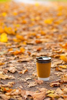 Walk with a cup of hot cocoa in the autumn park. Craft cup of coffee on the road with yellow fallen leaves.