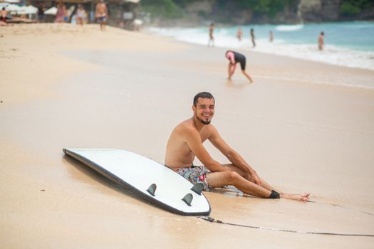 The guy is resting on a sandy tropical beach, after riding a surf. Healthy active lifestyle in summer vocation.