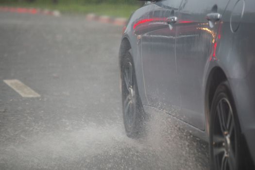 Rain water splash flow from wheels of silver car moving fast in daylight city with selective focus. Car moving on asphalt road during heavy summer storm rain.