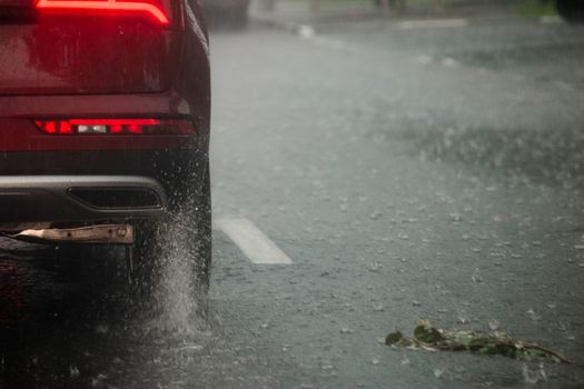Rain water splash flow from wheels of red car moving fast in daylight city with selective focus. Car moving on asphalt road during heavy summer storm rain. Torn green twig laying on the road.
