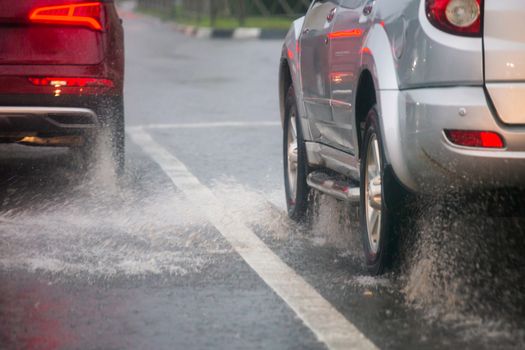 Rain water splash flow from wheels of silver and red cars moving fast in daylight city with selective focus. Car moving on asphalt road during heavy summer storm rain.