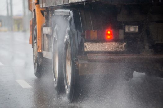 Rain water splash flow from wheels of heavy truck moving fast in daylight cit. Truck moving on asphalt road during heavy summer storm rain. Close-up with selective focus and blur.