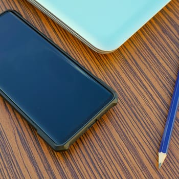 Computer notebook with black smartphone and a blue pencil on a wooden desk.