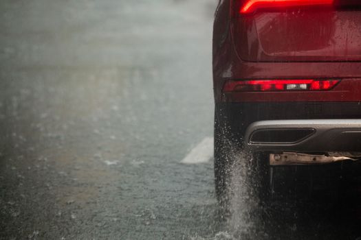 Rain water splash flow from wheels of red car moving fast in daylight city with selective focus. Car moving on asphalt road during heavy summer storm rain.