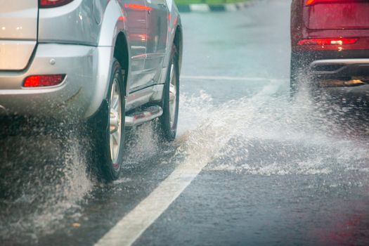 Rain water splash flow from wheels of silver and red cars moving fast in daylight city with selective focus. Car moving on asphalt road during heavy summer storm rain.