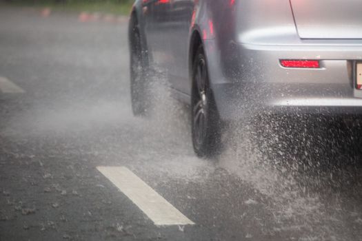 Rain water splash flow from wheels of silver car moving fast in daylight city with selective focus. Car moving on asphalt road during heavy summer storm rain.