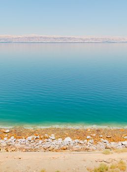 Vertical landscape view of the Dead sea shore, with salt crust and pebble beach near a tourist resort, and Israel territories mountain line on the horizon, from Madaba, Jordan, Middle East