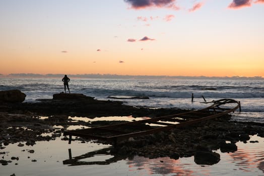 Silhouette of woman looking away at the Mediterranean sea, near an abandoned metal pier on a beach, at sunset,  in Batroun, Lebanon, Middel East