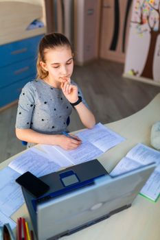 Beautiful young school girl working at home in her room with a laptop and class notes studying in a virtual class. Distance education and learning, e-learning, online learning concept during quarantine