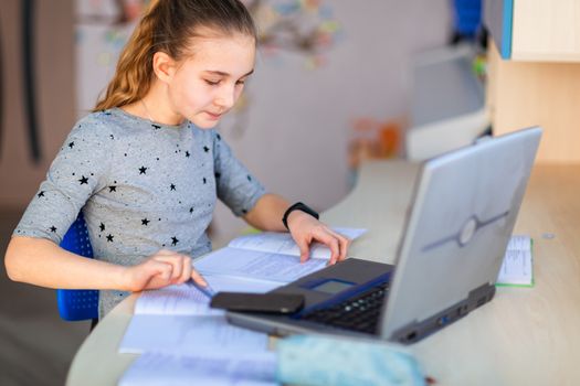 Beautiful young school girl working at home in her room with a laptop and class notes studying in a virtual class. Distance education and learning, e-learning, online learning concept during quarantine