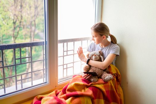 Sad little girl sitting on the window with a toy at home isolated, watching out. Coronavirus quarantine concept.