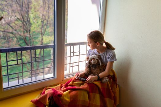 Sad little girl sitting on the window with a toy at home isolated, watching out. Coronavirus quarantine concept.