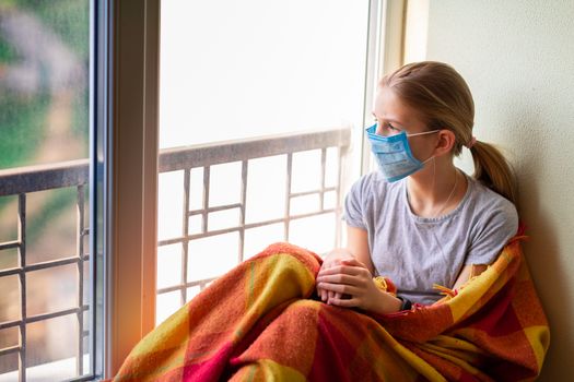 Sad little girl in protective mask sitting on the window at home isolated, watching out. Coronavirus quarantine concept.