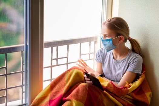 Sad little girl in protective mask sitting on the window with e-book or tablet reading and studying at home isolated. Coronavirus quarantine distance education concept.