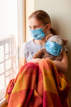 Sad little girl with teddy bear toy both in protective masks sitting on the window at home isolated, watching out. Coronavirus quarantine concept.