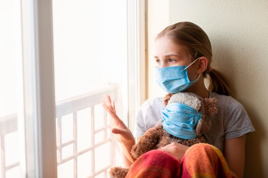 Sad little girl with teddy bear toy both in protective masks sitting on the window at home isolated, watching out. Coronavirus quarantine concept.