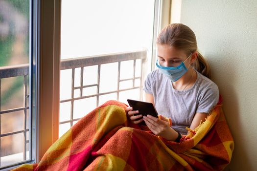 Sad little girl in protective mask sitting on the window with e-book or tablet reading and studying at home isolated. Coronavirus quarantine distance education concept.