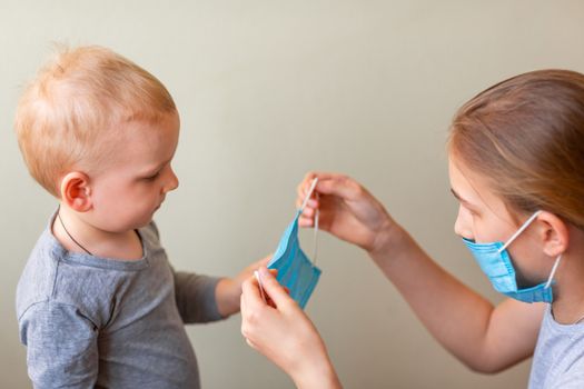 Teenage girl helping to his baby brother to wear a medical mask. Coronavirus protection, wearing masks concept