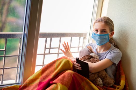 Sad little girl in protective mask sitting on the window with e-book or tablet reading and studying at home isolated. Coronavirus quarantine distance education concept.