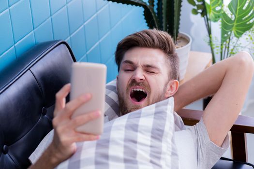 american man watching media content in a smart phone and yawn on a couch at home.