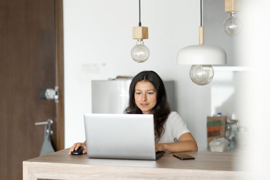 Beautiful young brunette girl working on a laptop at home in the kitchen