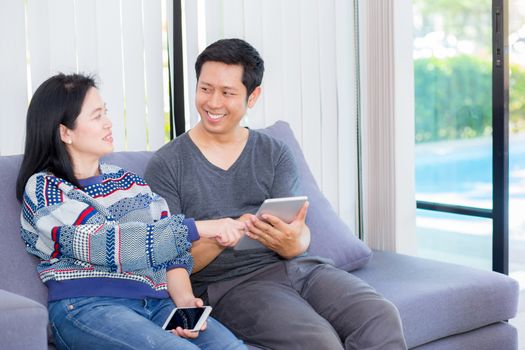 Two friends on line with multiple devices and talking sitting on a sofa in the living room in a house interior, communication concept.