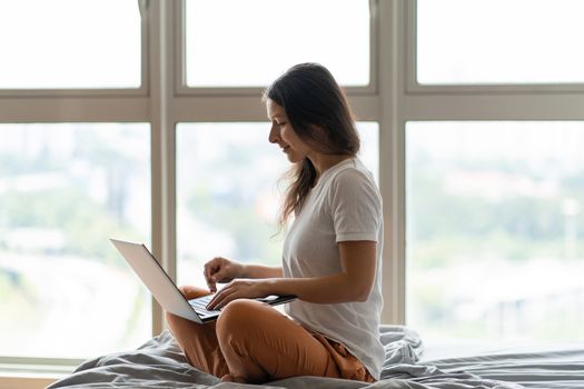 A beautiful young brunette girl is working on the laptop while sitting on a bed by a panoramic window with a beautiful view from a high floor. Stylish modern interior.Shopping on the Internet