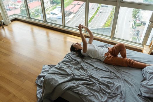 A beautiful young brunette girl is texting on the phone while lying on a bed by a panoramic window with a beautiful view from a high floor