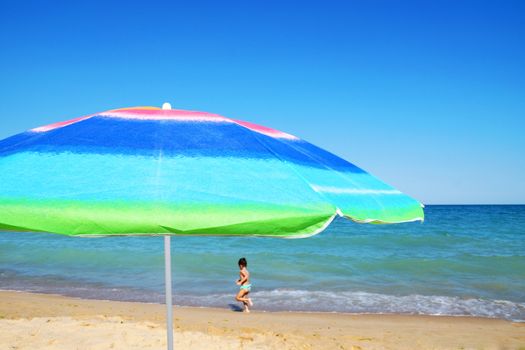 beach umbrella, pareo and sunglasses against the sea horizon and clear sky, child runs in the background, copy space