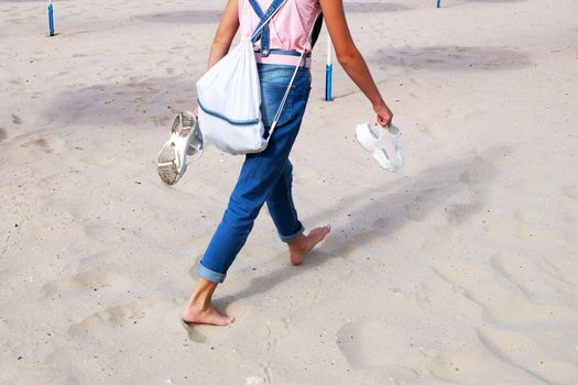 a girl in jeans walks on the sand barefoot, holding sandals in her hands, rear view