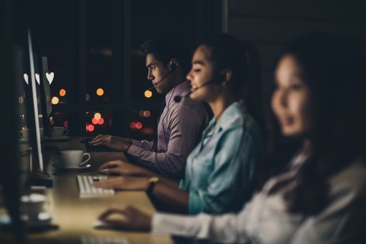 Asian male customer care service with business man smiling and working hard late in night shift at office,call center department,worker and overtime,7 days 24 hour,teamwork with colleagues for success