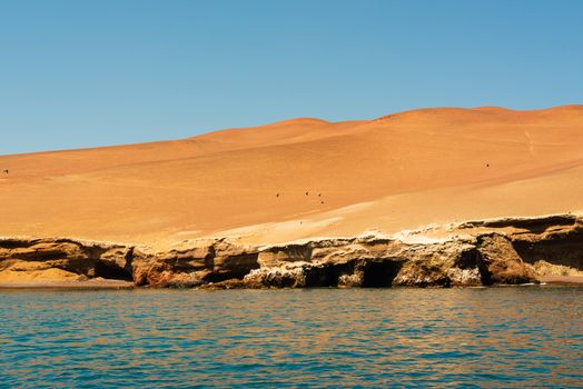 There is a blue sky over the Atacama desert with birds flying over it while the desert meets  the blue waters of the Pacific below.