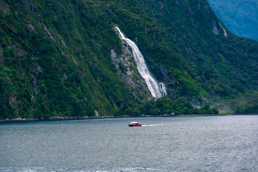 A red boat makes its way past a waterfall flowing into Milford Sound, New Zealand.