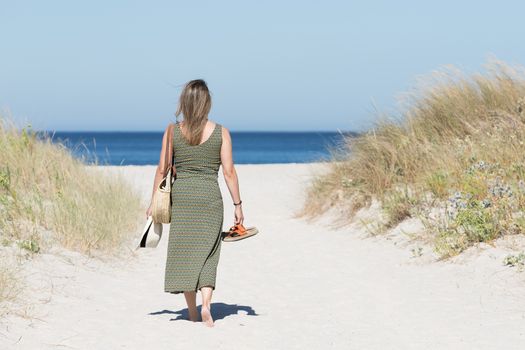 Middle-aged blonde woman in a dress walking barefoot along a sandy path towards the sea with her sandals in one hand and a hat in the other.