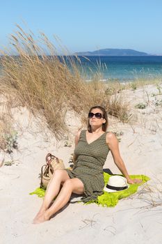 A middle-aged blonde woman with sunglasses sitting on the sand of a beach dune
