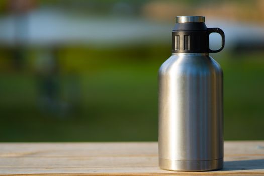 Large stainless steel thermos outside on a wooden table with a green summer blurred background while camping
