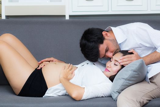 Husband kissing forehead of wife with pregnant belly of while sitting on sofa.