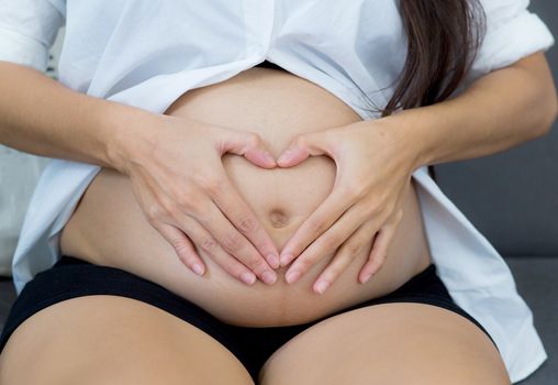 Pregnant asian woman holding gesture heart symbol shape belly with on sofa in home. Maternity concept.
