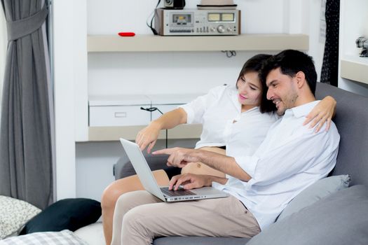 Portrait of a asian young couple husband and wife sitting on the sofa while using notebook computer in living room.