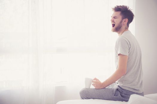 Lifestyle portrait of bedroom concept: Man holding a cup of coffee and yawn with on bedroom good morning.