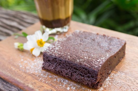 Chocolate Brownie Cake and Latte Coffee and Daisy Flower on Chopping Board or Cutting Board on Wood Table on Green Tree Background.