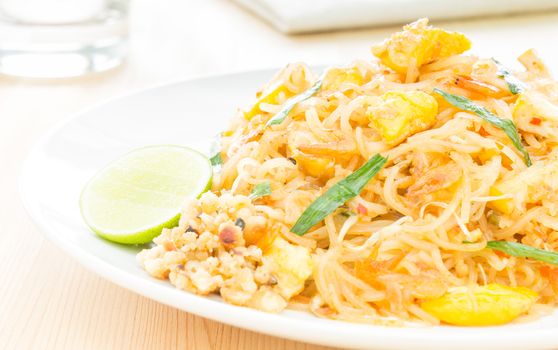 Pad Thai or Thai fried noodles on white dish on wood table. Glass and napkin egg dry shrimps onions sprouts lemon peanut