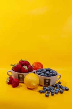  strawberriesand blueberries in ceramic cups next to one lemon and tangerine shot against a yellow background