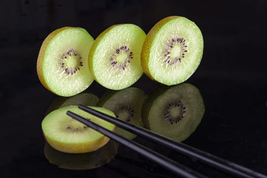 Kiwi one piece sliced and three pieces chopped stand and next to Chinese sticks on a black background