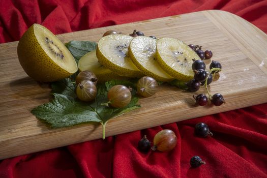 Kiwi sliced, gooseberries and black currants with leaves on a wooden board on a  red background