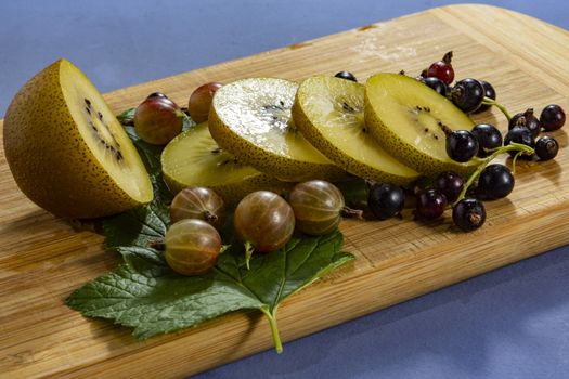 Kiwi sliced, gooseberries and black currants with leaves on a wooden board on a blue  background