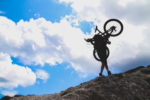 sport man in action lifting bike on his shoulder on rock mountain with blue sky background.