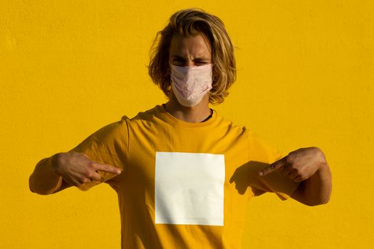 Blond young man with mask pointing at shirt on a yellow background. Summer coronavirus concept