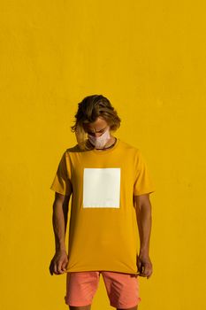 Blond young man with mask on a yellow background looking at the shirt. Summer coronavirus concept